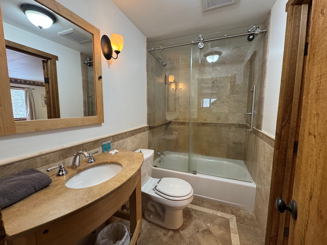 Bathroom 2: Stone-tile tub with sliding glass shower door, curved stone vanity, wood mirror