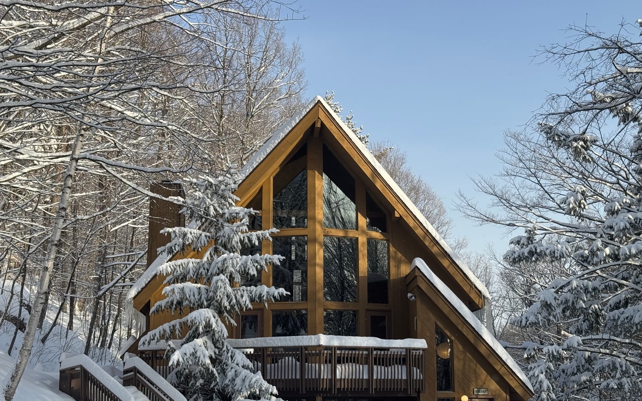 The cedar A-frame in deep snow: a two-story glass gable, a snow-laden spruce and the front deck under a blue sky.